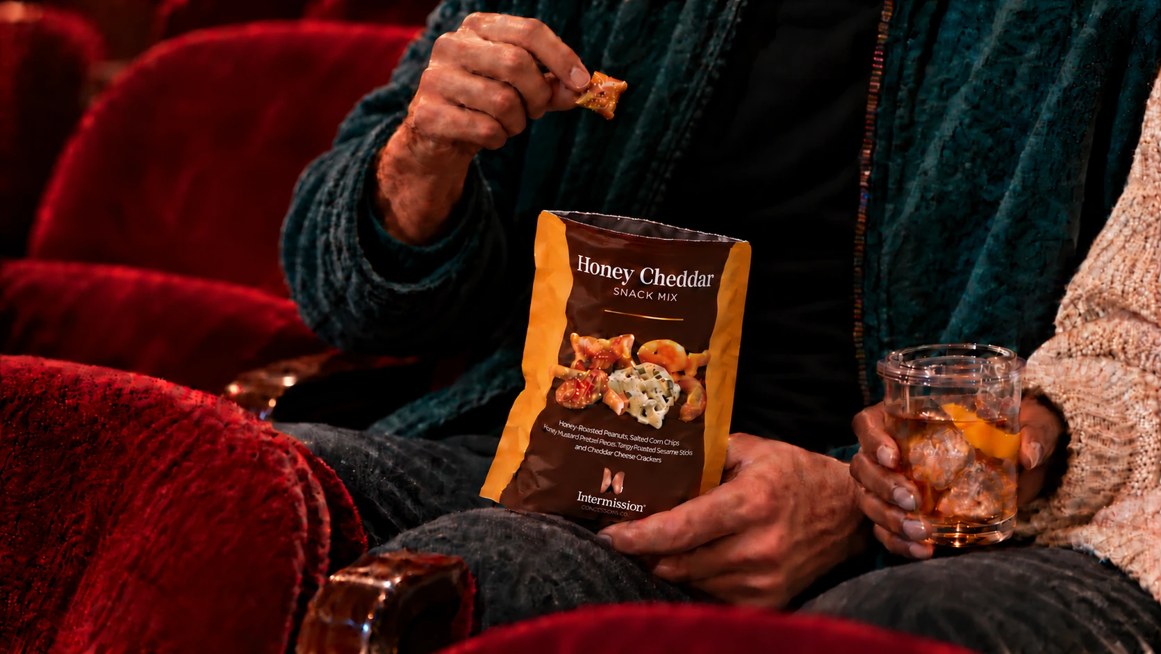 A man's hand holding a pretzel between his thumb and finger above an open Intermission Honey Cheddar snack mix pouch while a woman holds a cocktail in a clear lidded theater cup beside him
